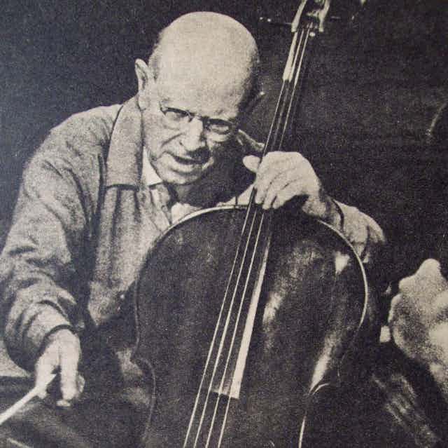Un hombre calvo sentado sujeta un violonchelo con la mano izquierda y el arco con la mano derecha