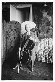 Un hombre joven sentado en una silla sujeta un violonchelo y un arco y mira de lado fuera del encuadre.
