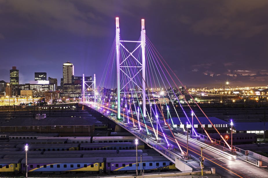 Cityscape by night, with a bridge in the foreground, spanning rows of trains on a railway line