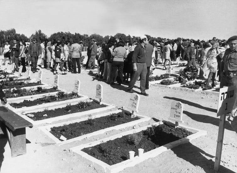 People are seen walking around small gravestones in a row, in front of fresh, long plots of dirt in a black and white photo.