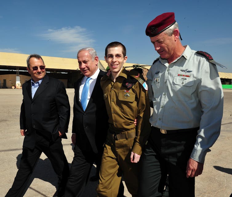 Gilad Shalit and Benjamin Netanyahu walk with two other men in suits on an airplane tarmac.