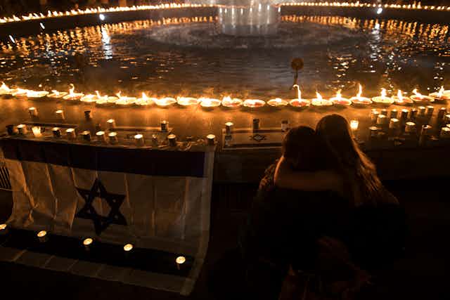 People embrace next to an Israeli flag and a large circle of candles around a circular fountain or pool of water at nighttime.