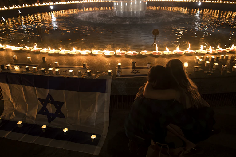 People embrace next to an Israeli flag and a large circle of candles around a circular fountain or pool of water at nighttime.