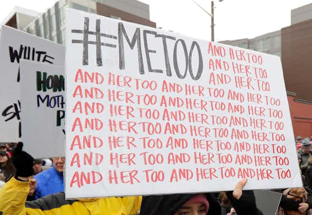 People are barely visible behind large white signs they are holding, the first of which says '#MeToo and her too' over and over.