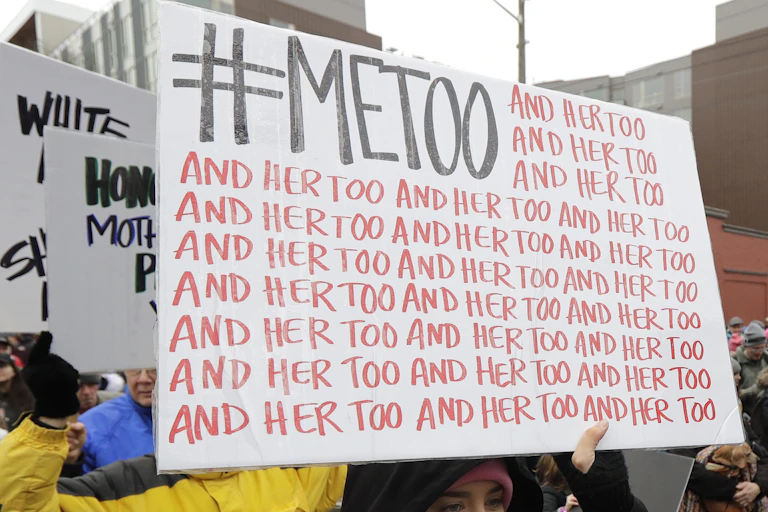 People are barely visible behind large white signs they are holding, the first of which says '#MeToo and her too' over and over.