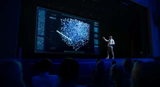 a man stands on a stage and a screen behind him shows a scattergraph