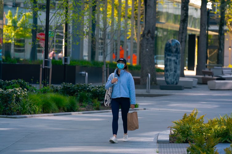 a woman wearing a surgical mask and carrying bags walks down a street