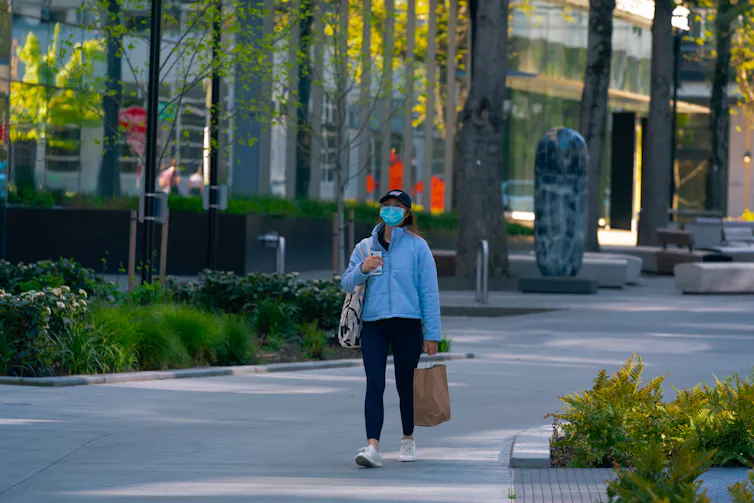a woman wearing a surgical mask and carrying bags walks down a street