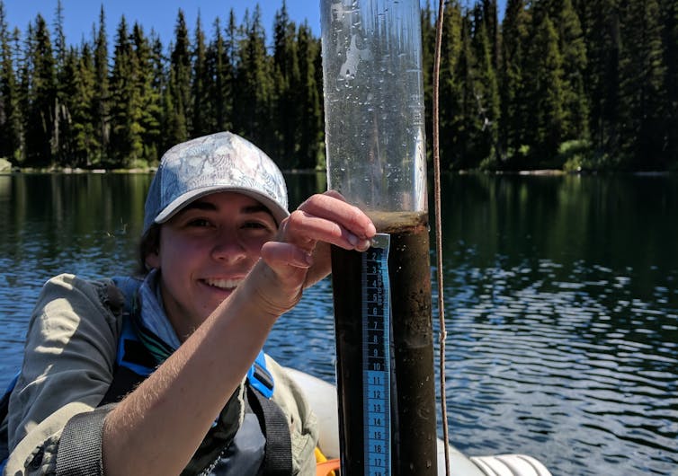 A woman sitting an inflatable boat, wearing a life jacket, holds a long tube filed with lake bottom sediment.