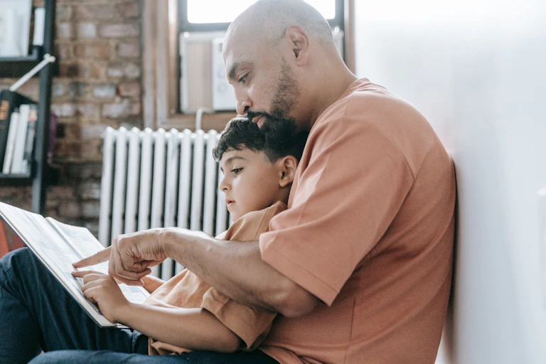 A man sits with a young boy, reading a book.