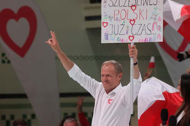 Donald Tusk giving a peace sign and waving a placard that says "it's time for a happy Poland" in Polish.