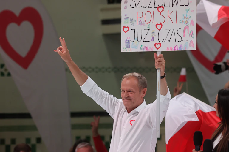 Donald Tusk giving a peace sign and waving a placard that says "it's time for a happy Poland" in Polish.