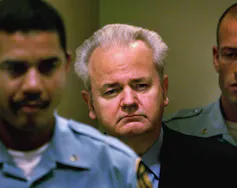 A round-faced man with grey hair is escorted into a courtroom between two court officers.