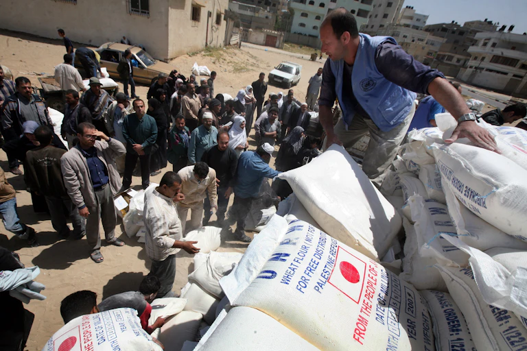 Man unloads a pile of large white sacks off, surrounded by a crowd of men