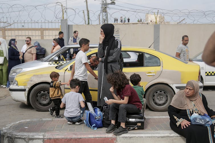 Uma família espera com sua bagagem ao lado de táxis estacionados em frente a um muro com arame farpado.