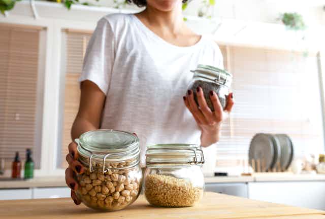 A woman wearing a white shirt holds two glass jars, one with chickpeas and one with black rice. A jar of pine nuts sits on the wooden countertop.