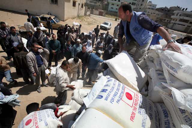 Homem descarrega uma pilha de grandes sacos brancos, cercado por uma multidão de homens
