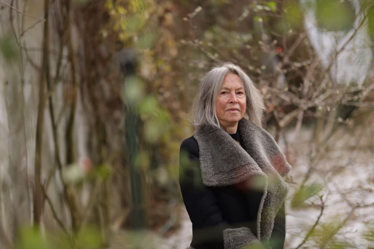 Louise Glück wearing a thick jacket, standing outside surrounded by tree branches