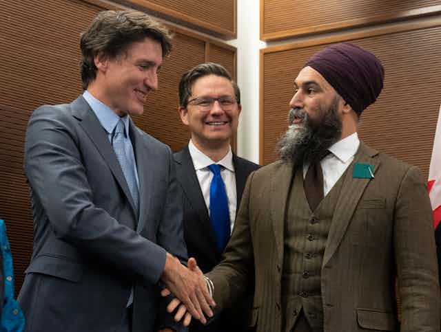 Two men shakes hands, one wearing a turban, while another man smiles between them.