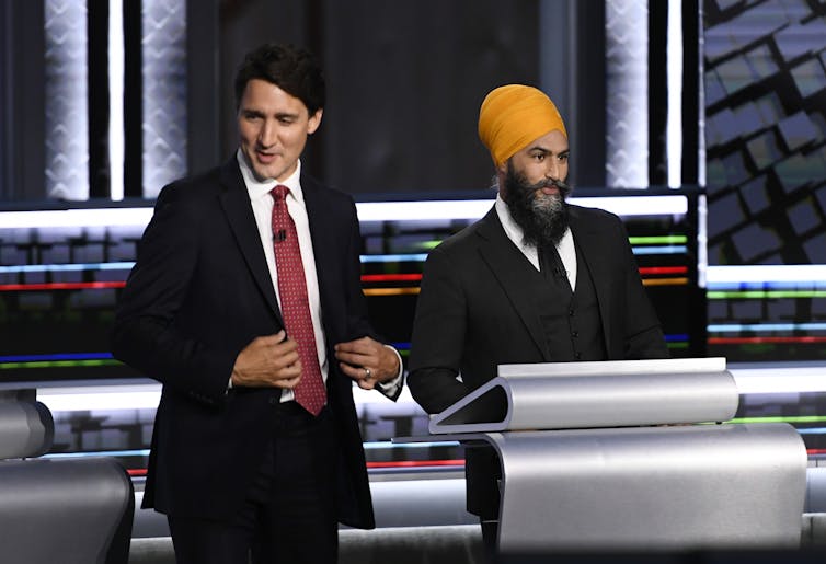 A man with dark hair in a dark suit stands next to another man in a suit donning an orange turban.
