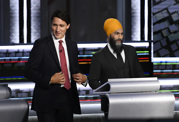 A man with dark hair in a dark suit stands next to another man in a suit donning an orange turban.