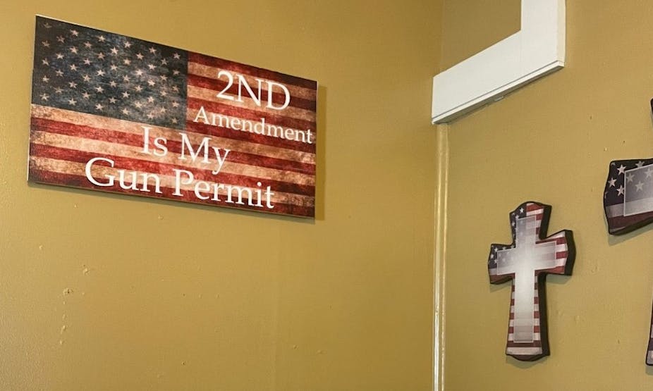A brown-yellow wall features a flag decoration that says '2nd Amendment Is My Gun Permit' and two crosses in the colors of the U.S. flag.