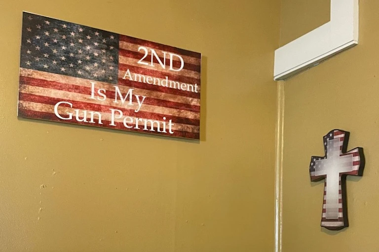 A brown-yellow wall features a flag decoration that says '2nd Amendment Is My Gun Permit' and two crosses in the colors of the U.S. flag.