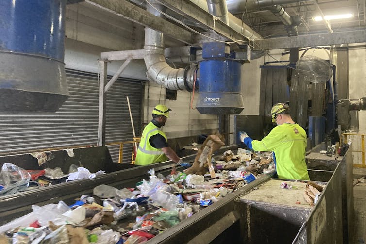 Two workers, in bright yellow, stand at a conveyor belt covered in plastics in a recycling facility.