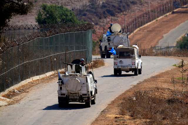 White armoured vehicles with UN flags and solidiers along a border road. 