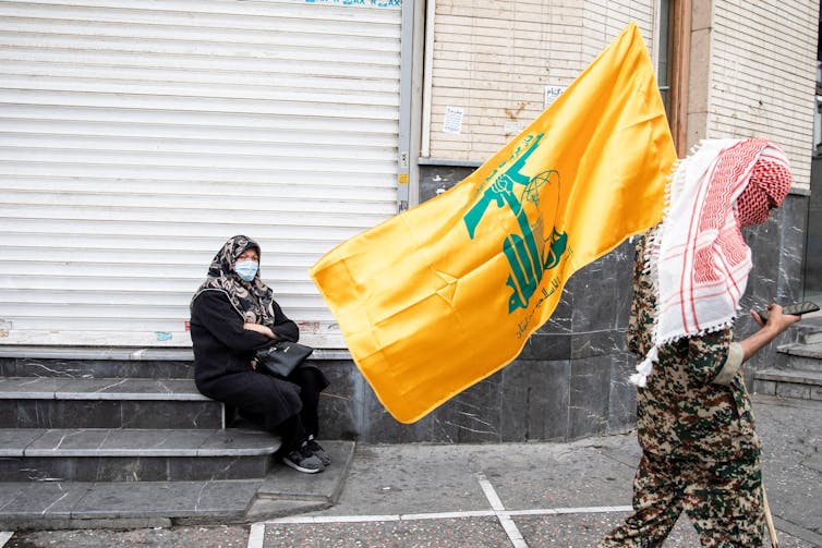 Un homme vêtu d’un kefiyeh rouge et d’une tenue militaire tient le drapeau jaune du Hezbollah alors qu’il marche dans une rue devant une femme assise portant un foulard et une abaya noire.