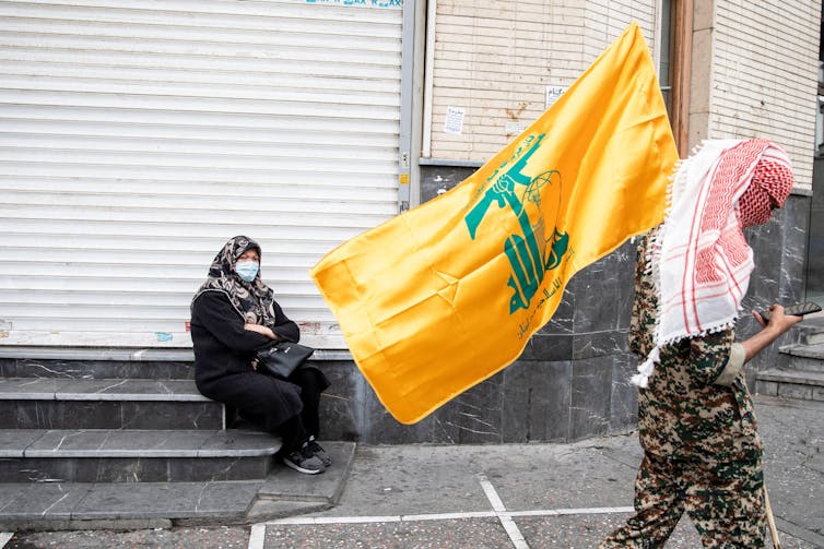 A man in a red kefiyeh and military garb holds the yellow Hezbollah flag as he walks down a street past a seated woman in a headscarf and black abaya.