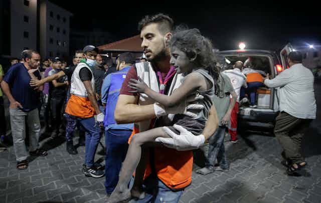 A Palestinian man carries a young girl in his arms with an ambulance in the background.