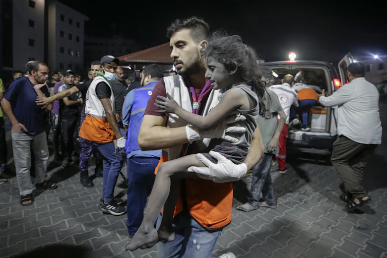 A Palestinian man carries a young girl in his arms with an ambulance in the background.