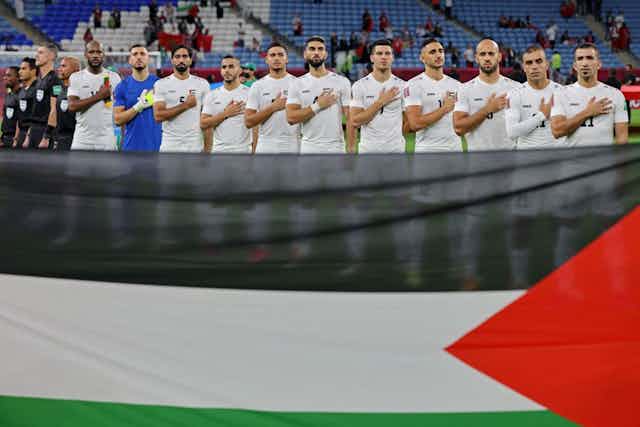 A row of men in white sports kit, with their right hands over their hearts; flag in the foreground in black, white, green and red