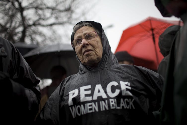 An older woman with glasses wears a black windbreaker that reads Peace, Women in Black.