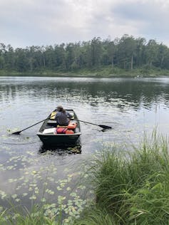 rowboat with one woman in it on a lake with woodsy shoreline