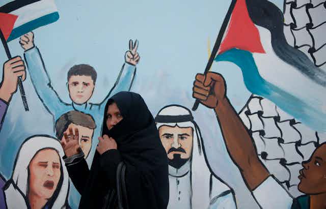 A woman in a headscarf walks past a mural of people flashing 'V' peace signs and holding Palestinian flags.