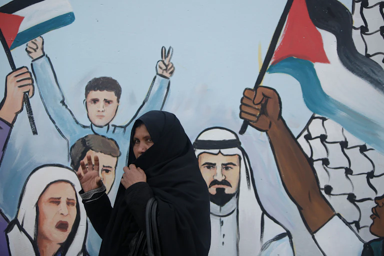 A woman in a headscarf walks past a mural of people flashing 'V' peace signs and holding Palestinian flags.