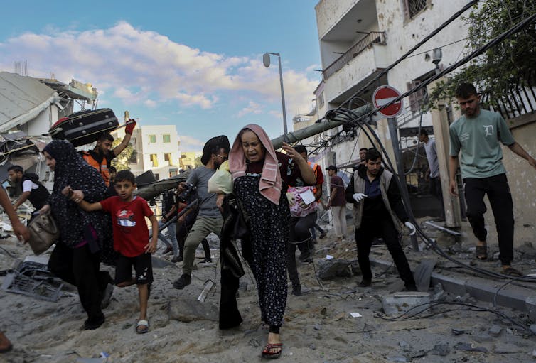 Several people, including women and children, running out of their homes. Behind them are some partially damaged buildings.