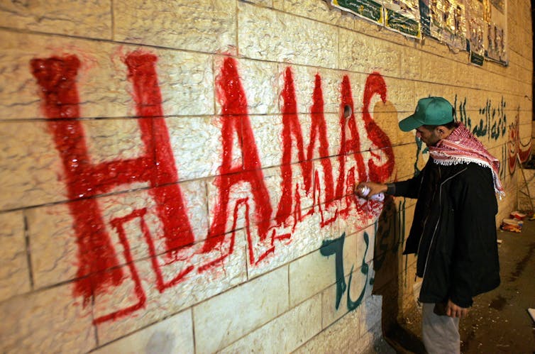 A man in a cap paints the word Hamas in large letters on a wall.