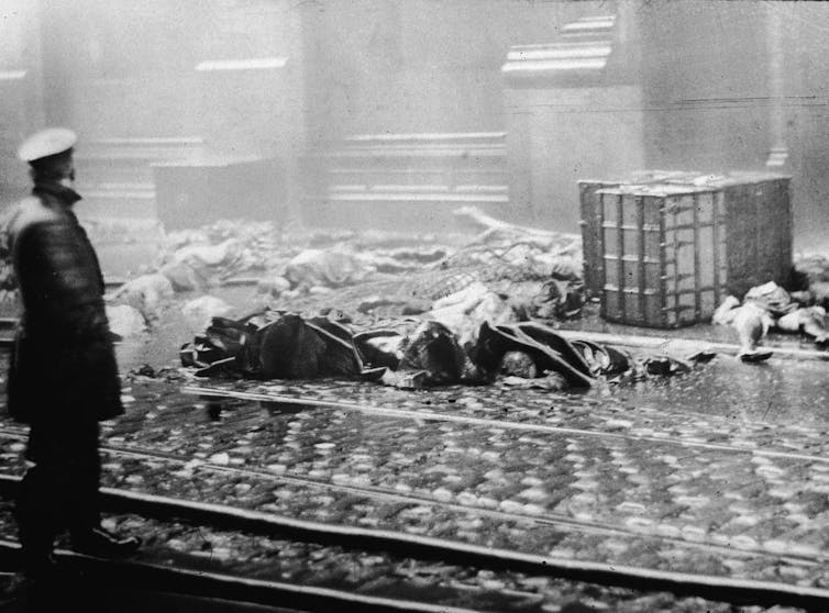 A black and white photo of a man looking from a few feet away at dead bodies crumpled on a sidewalk.