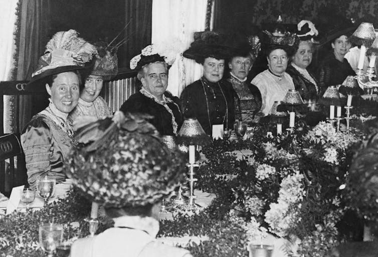 A black and white photo of formally dressed women around a dining table decorated with plants and candles.
