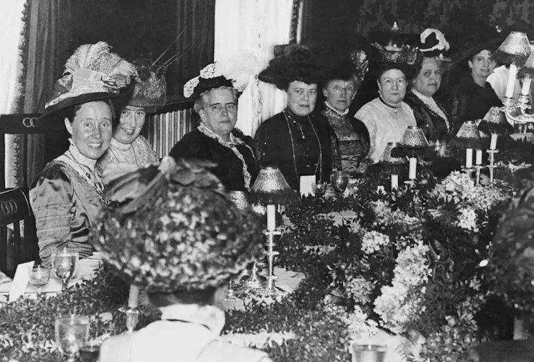 A black and white photo of formally dressed women around a dining table decorated with plants and candles.