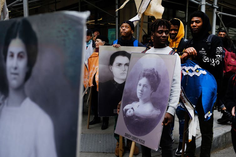 Young men hold posters printed with black and white photographs of women as they stand on a city street.