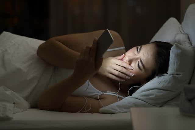 Young woman looking at brightly lit smartphone in bed, hand to face, looking worried or shocked