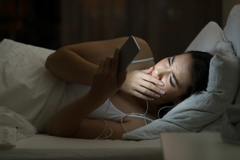 Young woman looking at brightly lit smartphone in bed, hand to face, looking worried or shocked