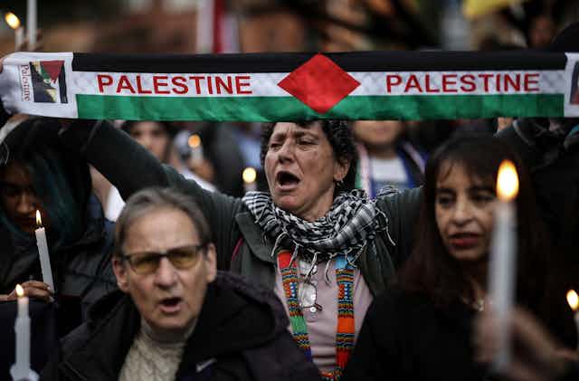 A woman at a demonstration holds a scarf in the colours of the Palestinian flag with the word Palestine on it.