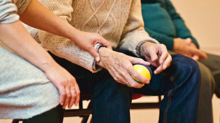 An elderly man holds a stress ball, while another person helps.