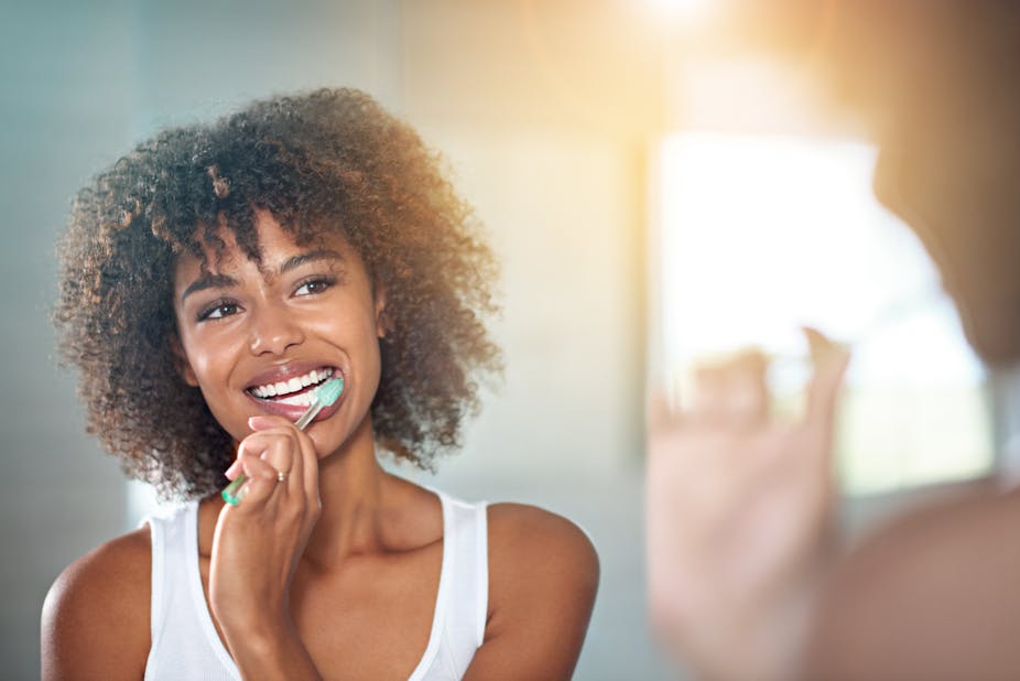 On voit l'image dans son miroir d'une jeune femme souriante en train de se brosser les dents.