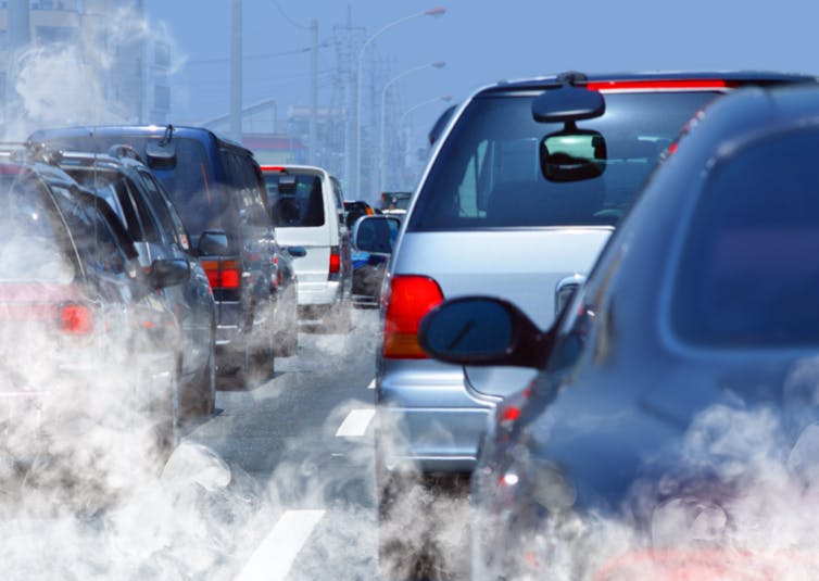 A line of cars waiting in traffic emitting exhaust fumes.